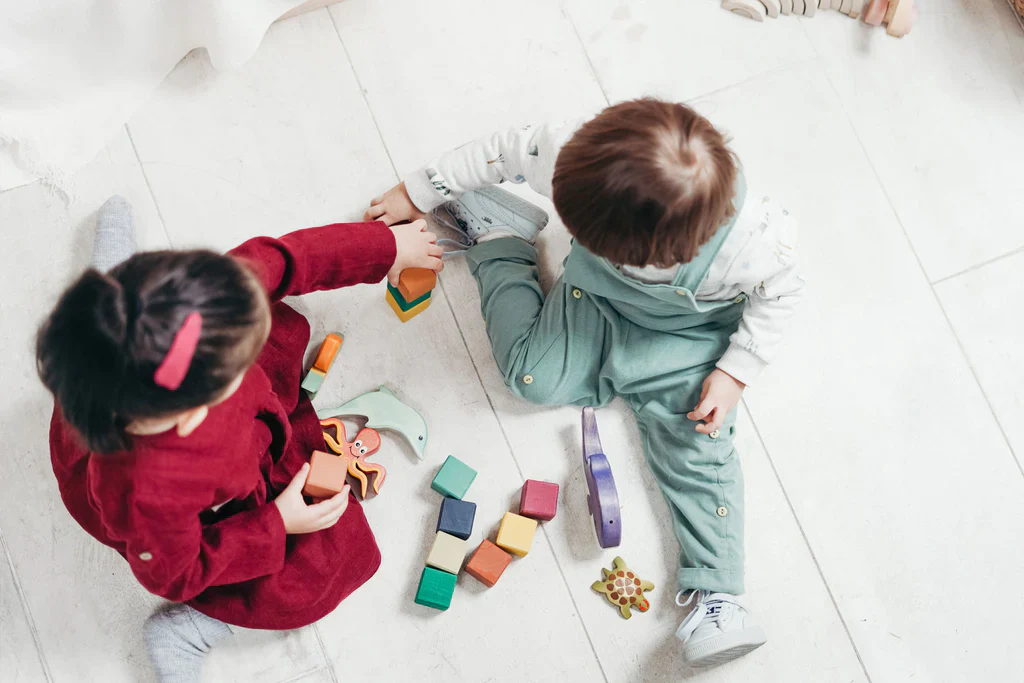 Two toddlers playing with wooden blocks and animal figures on a light wooden floor