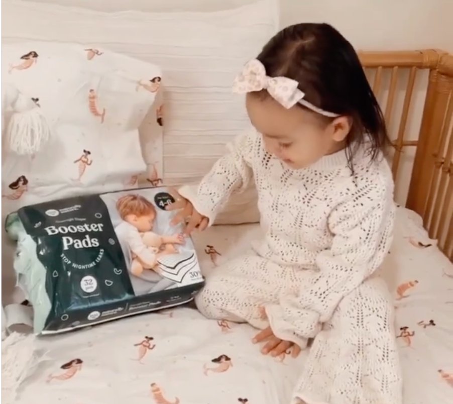A little girl in a white knit dress holding a Naturally Nature Booster Pads pack on a bed with mermaid-print sheets